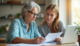 An older woman and a younger woman sit together at a table, reviewing paperwork with a laptop open nearby. The older woman writes on a document while the younger woman looks on supportively, creating a warm, collaborative atmosphere.